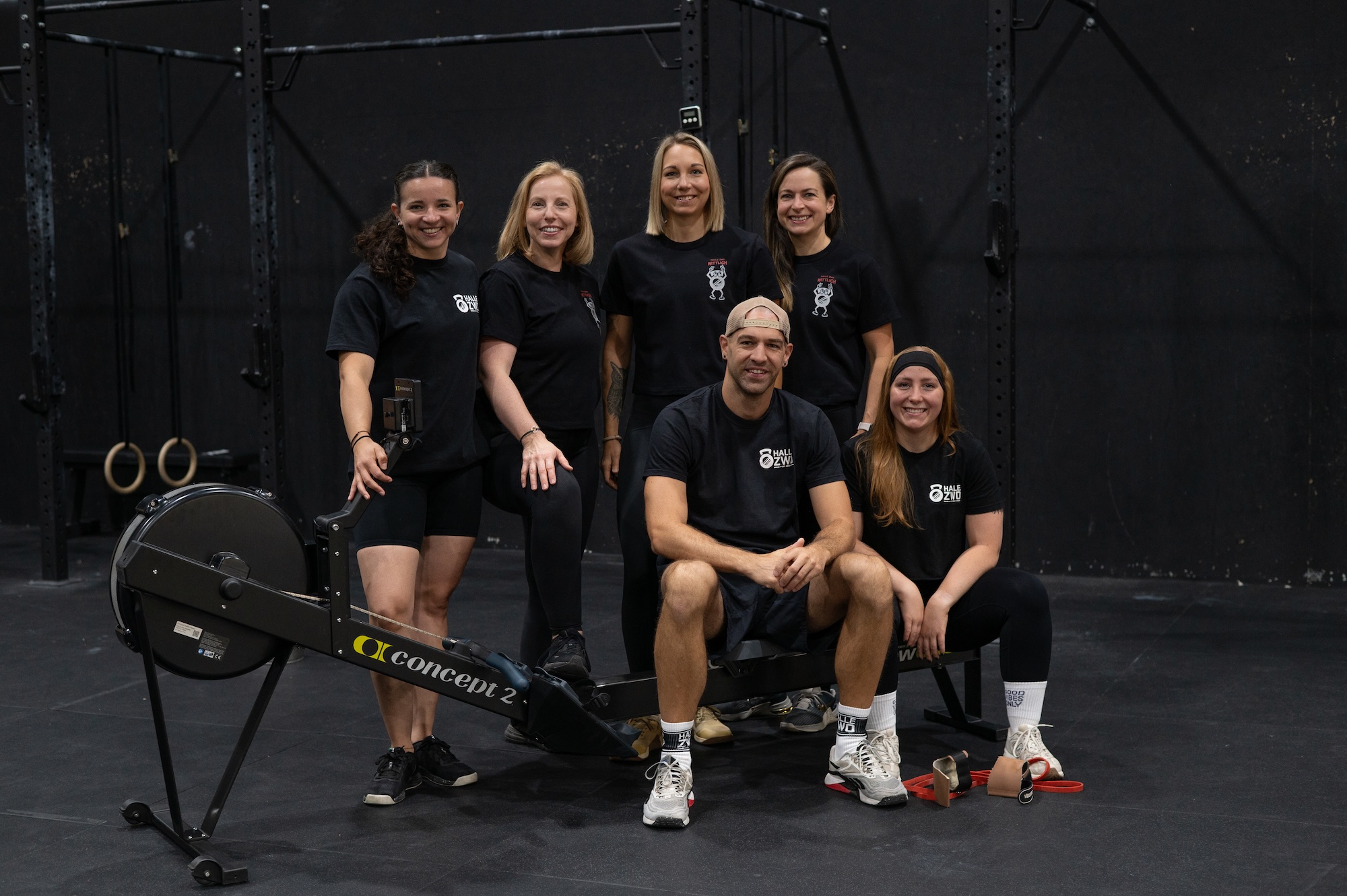 Das Halle Zwo Trainerteam - Sechs engagierte Fitness-Coaches in schwarzen Halle Zwo T-Shirts posieren gemeinsam am Concept2 Rudergerät in der CrossFit Box